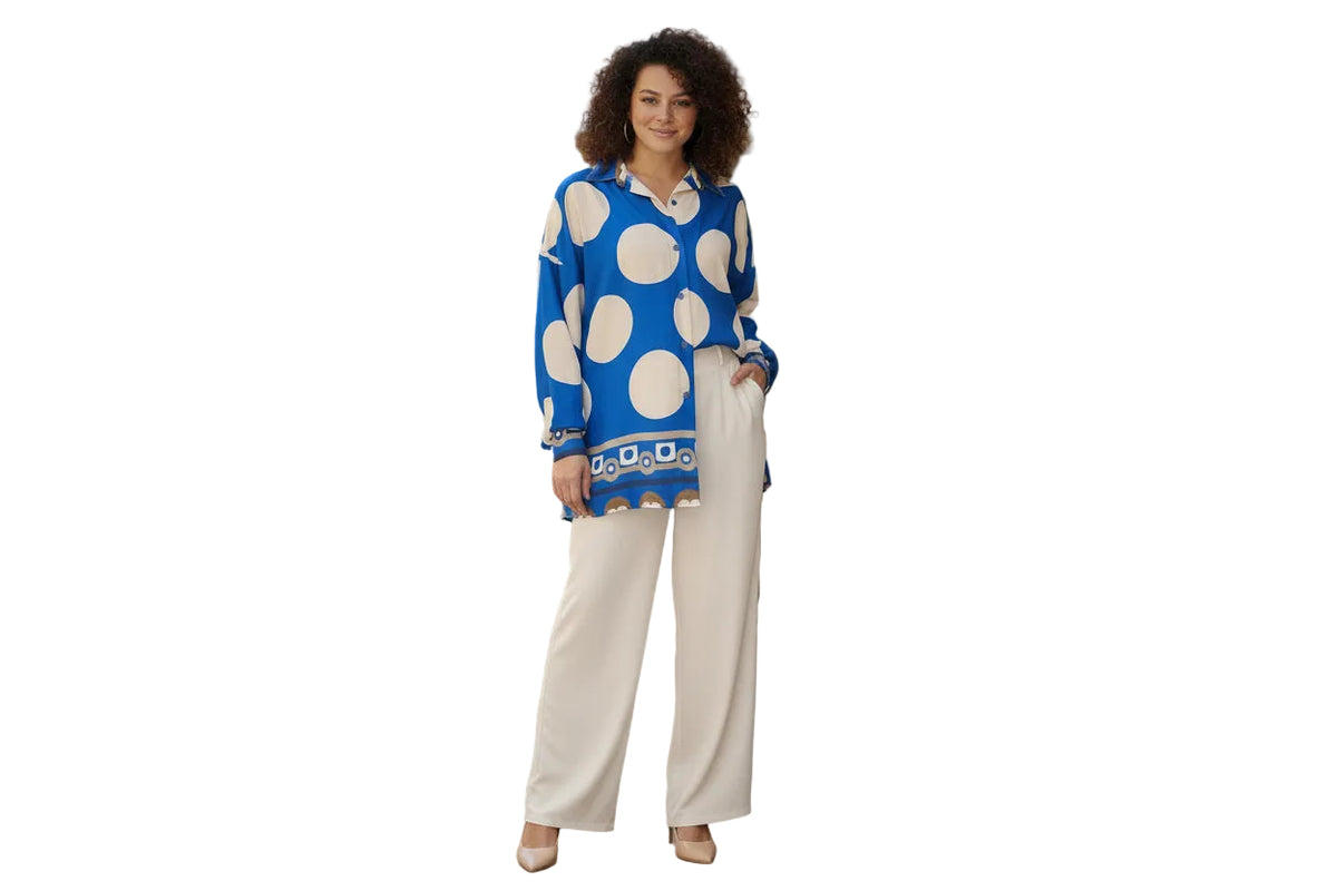Woman wearing a blue polka dot shirt and white pants standing in an outdoor setting with tables and chairs.